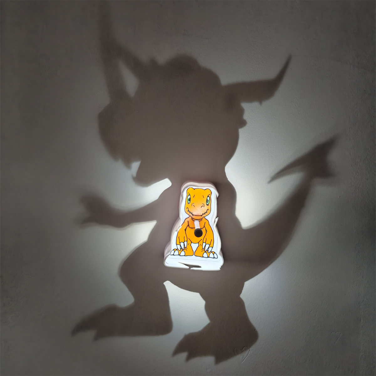 Agumon Digimon-inspired shadow lamp casting a gigantic dinosaur monster silhouette on the wall around its glowing orange character panel