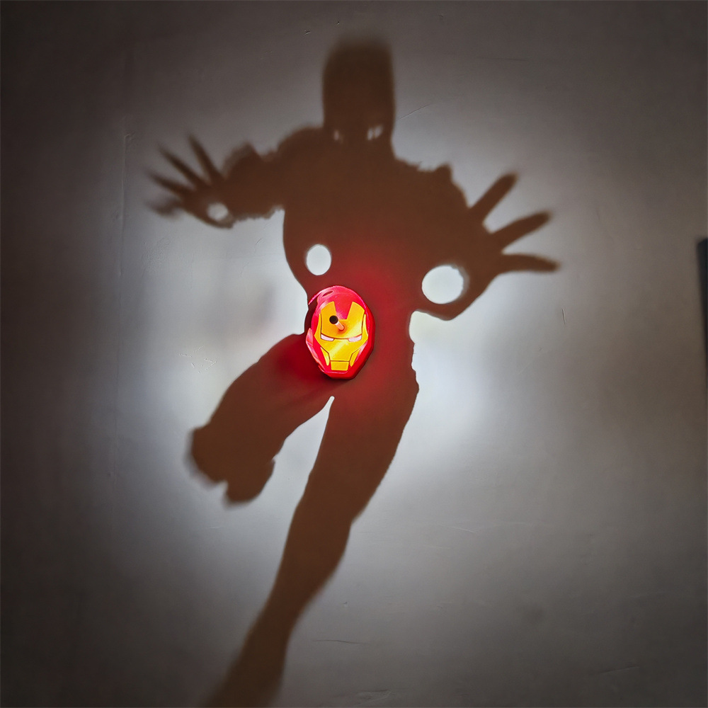 Iron Man-inspired shadow projection lamp with illuminated red and gold mask casting large armored superhero shadow with outstretched hands on a wall